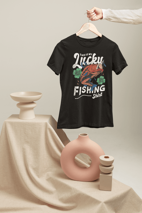 Black Lucky Fishing T-Shirt displayed on a hanger with abstract ceramic decor in the background